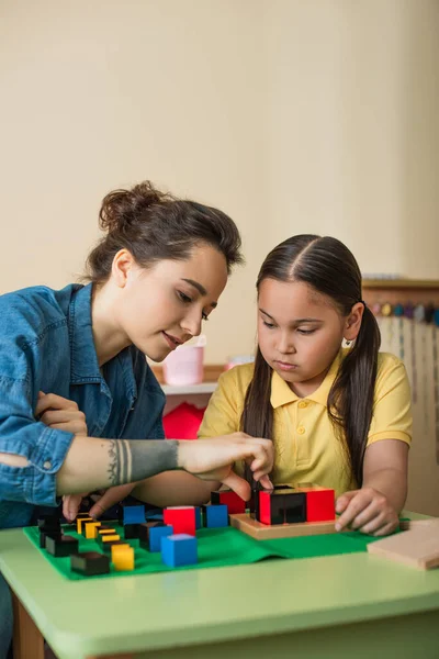 Mujer tatuada jugando bloques multicolores juego con chica asiática en la escuela montessori - foto de stock