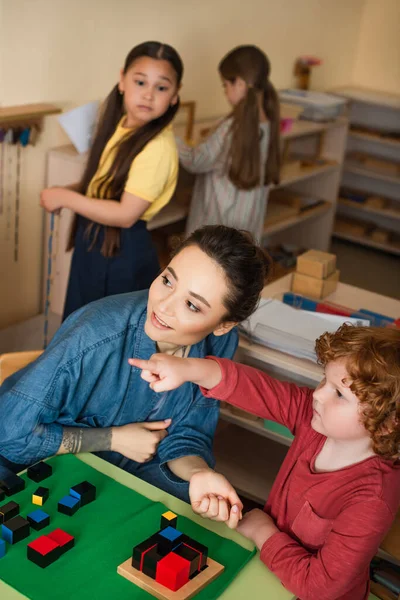 Chico apuntando con el dedo mientras juega cubos de madera juego cerca de profesor y borrosa interracial niñas - foto de stock