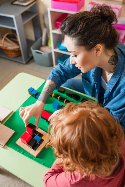 Vista ad alto angolo di giovane insegnante e rossa bambino che gioca con cubi colorati nella scuola montessori — Foto stock