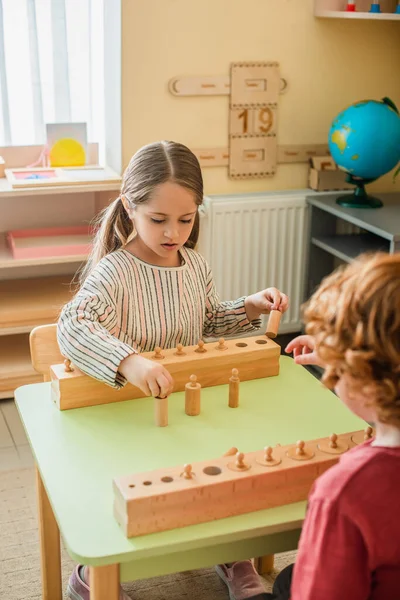 Дівчина грає в гру з дерев'яним елементом біля розмитої дитини в школі Монтессорі — стокове фото