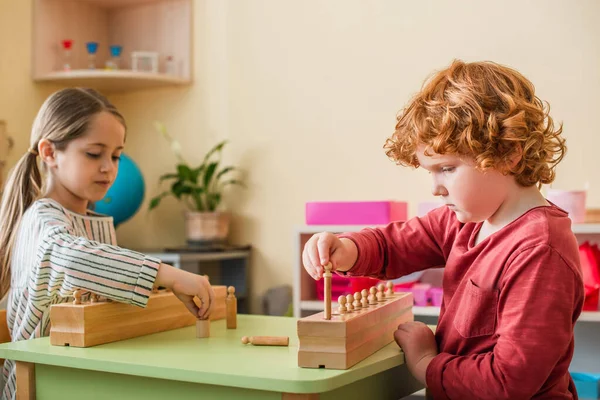 Menino encaracolado jogando jogo com elementos de madeira perto da menina na escola montessori — Fotografia de Stock