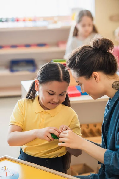 Junge Lehrerin hilft asiatischem Mädchen bei der Kombination von Puzzle in der Nähe von Mädchen auf verschwommenem Hintergrund — Stockfoto