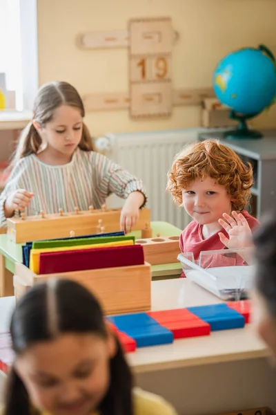 Niño saludando de la mano mientras llama borrosa profesor cerca de las niñas en el aula - foto de stock