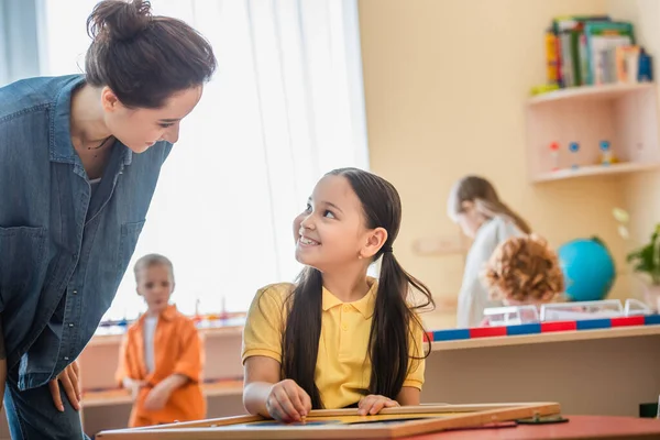 Cheerful asian girl combining puzzle near teacher and kids on blurred background — Stock Photo