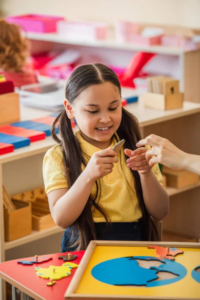 Cheerful asian girl holding puzzle element near teacher in montessori school — Stock Photo