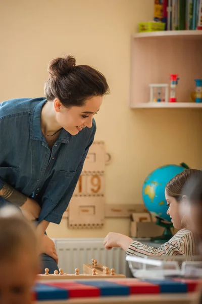 Smiling teacher looking at playing girl near boy on blurred foreground — Stock Photo
