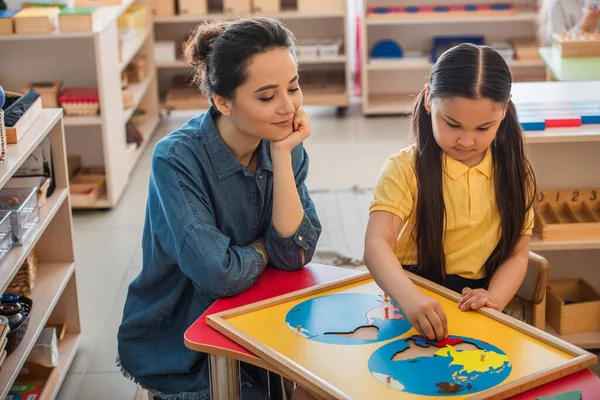 Молодой учитель глядя на азиатского ребенка, объединяющего карту земли головоломка в Монтессори школы — стоковое фото