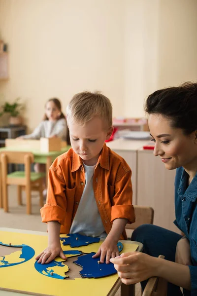 Мальчик сочетает карту земли головоломка рядом улыбающийся учитель и девочка на размытом фоне — стоковое фото