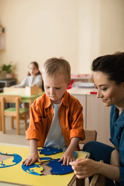 Boy combining earth map puzzle near smiling teacher and girl on blurred background — Stock Photo