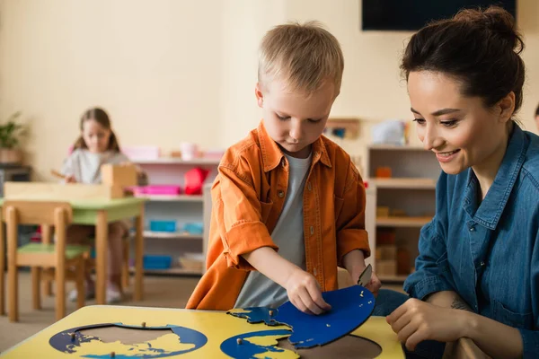 Boy combining earth map puzzle near smiling teacher and girl on blurred background — Stock Photo