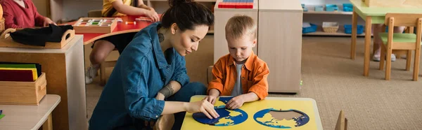 Junge Lehrerin hilft Junge beim Kombinieren von Erdkartenpuzzle, Banner — Stockfoto