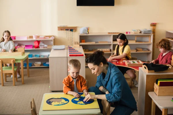 Professor com menino combinando quebra-cabeça mapa terra perto de crianças jogando jogos educativos — Fotografia de Stock