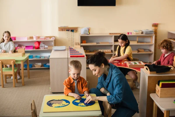 Insegnante con ragazzo combinando puzzle mappa della terra vicino ai bambini che giocano giochi educativi — Foto stock