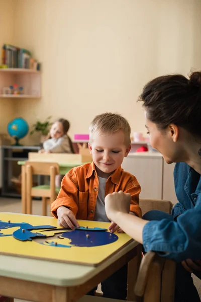 Cheerful boy combining earth map puzzle near teacher and blurred girl — Stock Photo