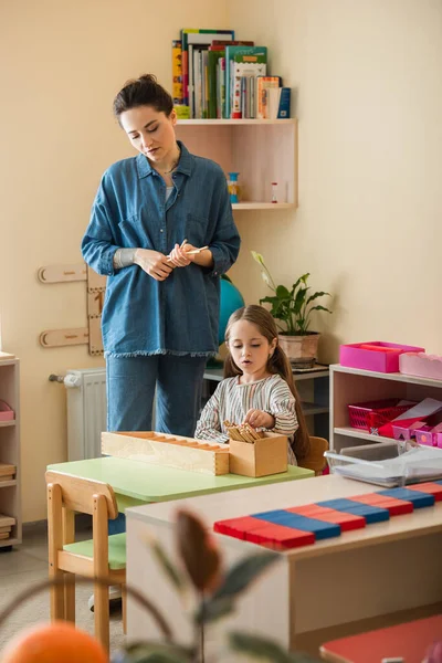 Young teacher standing near girl playing educational game in montessori school — Stock Photo