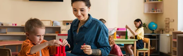 Teacher smiling near boy making tower from wooden cylinders in montessori school, banner — Stock Photo