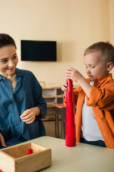 Young teacher smiling near boy making tower from red wooden cylinders — Stock Photo