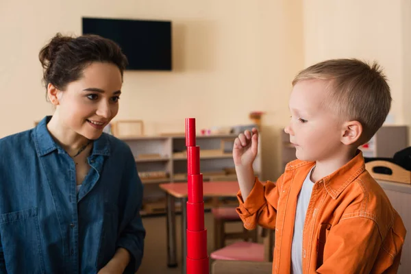 Joven profesor sonriendo cerca de niño y torre hecha de cilindros rojos - foto de stock