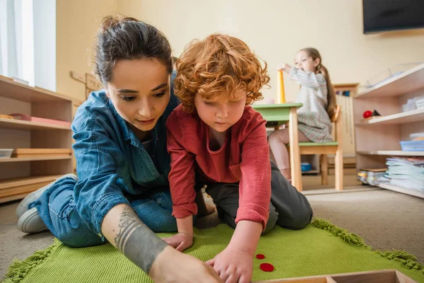 Tattooed teacher playing on floor with redhead boy near girl on blurred background — Stock Photo