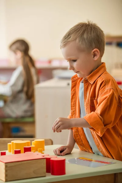 Chico jugando con rojo y amarillo cilindros cerca borrosa chica en montessori escuela - foto de stock