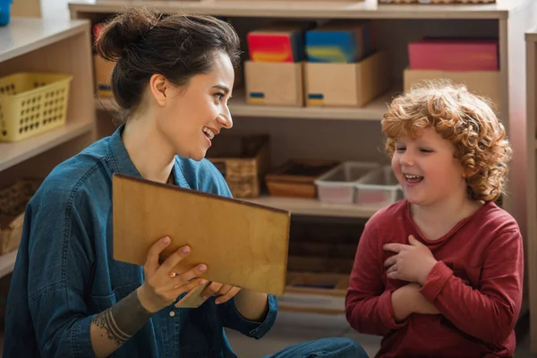 Happy teacher showing wooden board while playing educational game with curly boy — Stock Photo