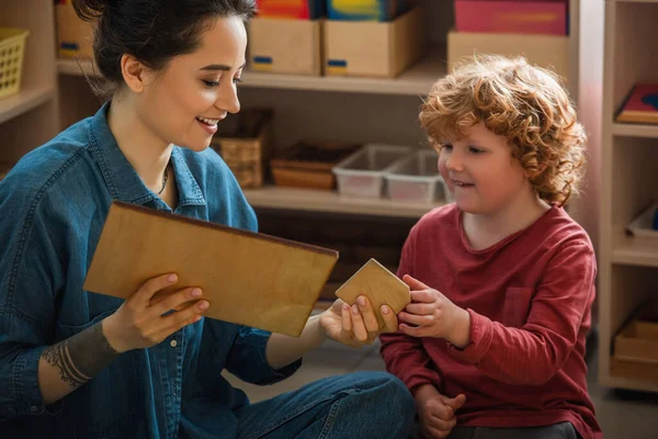 Insegnante sorridente che tiene tavole di legno mentre gioca con il ragazzo rossa nella scuola montessori — Foto stock