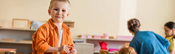 Веселий хлопчик, посміхаючись на камеру біля розмитого вчителя та дітей у школі Монтессорі, банер — стокове фото