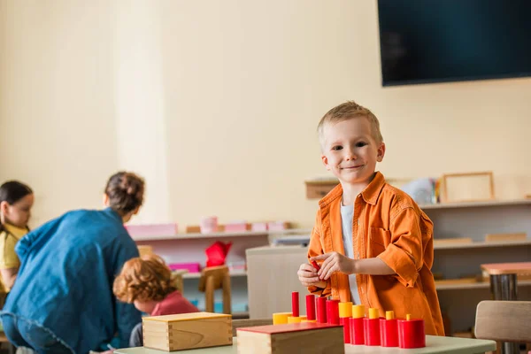 Cheerful boy looking at camera near tower made from red cylinders and blurred teacher with interracial girls — Stock Photo