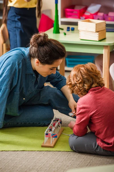 Redhead boy playing with small international flags near teacher on floor in montessori school — Stock Photo