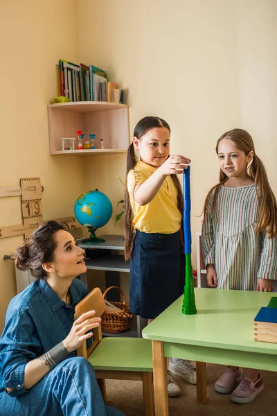 Giovane insegnante guardando ragazze interrazziale che fanno torre da cilindri di legno — Foto stock