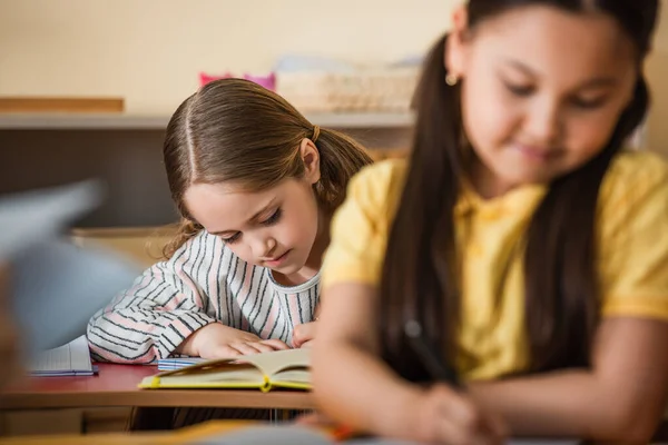 Chica escritura durante lección cerca asiático amigo en borrosa primer plano - foto de stock