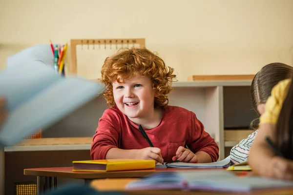 Alegre chico escribiendo en cuaderno cerca borrosa profesor y niñas en el aula - foto de stock