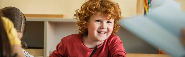 Pelirrojo chico sonriendo cerca de chica en montessori escuela en borrosa primer plano, bandera - foto de stock