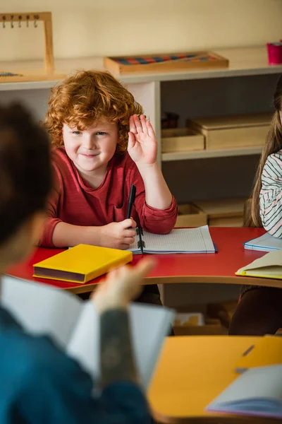 Rousse garçon levant la main près flou enseignant dans montessori école — Photo de stock