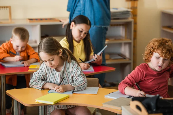 Niños multiétnicos que escriben en cuadernos cerca del maestro sobre fondo borroso - foto de stock