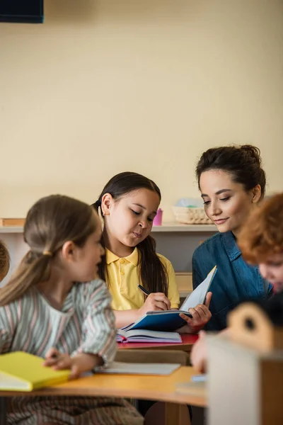 Asiatico ragazza scrittura in notebook vicino insegnante e bambini su sfocato primo piano — Foto stock