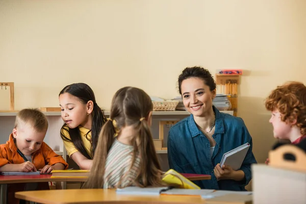 Joven profesor con libro sonriendo cerca interracial niños en montessori aula - foto de stock