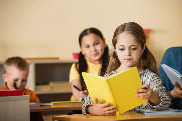 Fille lecture livre pendant leson dans montessori école près flou multiethnique amis — Photo de stock
