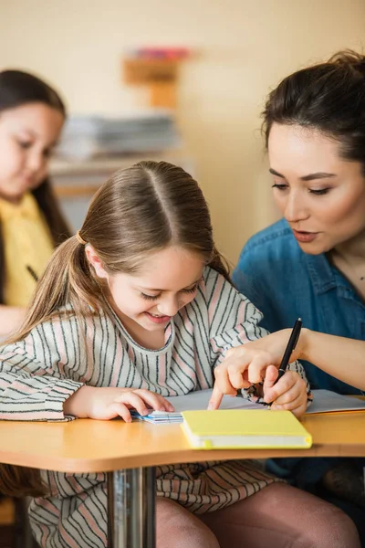 Joven profesor apuntando cerca sonriente niño escritura en notebook y asiático chica en borrosa fondo - foto de stock