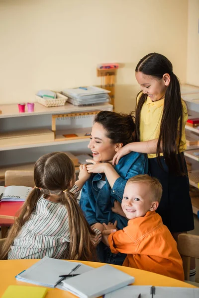Excited multicultural kids embracing happy teacher in classroom — Stock Photo