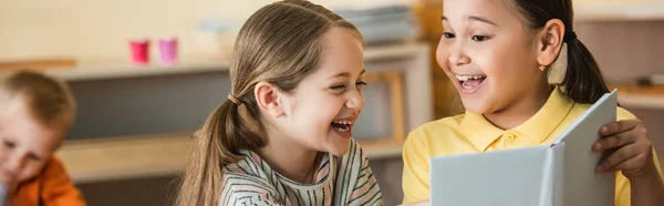 Fröhliche interrassische Mädchen, die Buch in der Nähe von Jungen auf verschwommenem Hintergrund betrachten, Banner — Stockfoto