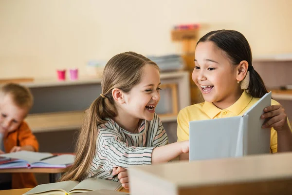 Excité fille pointant à livre près de joyeux asiatique ami dans montessori école — Photo de stock