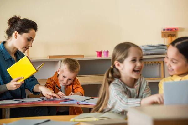 Professor apontando para notebook perto de escrever menino enquanto meninas inter-raciais rindo em primeiro plano turvo — Fotografia de Stock