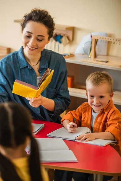 Sonriente profesor sosteniendo libro cerca feliz chico y borrosa chica en montessori escuela - foto de stock