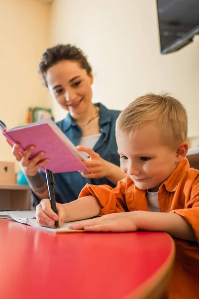 Ragazzo concentrato scrittura dettatura vicino insegnante offuscata con libro — Foto stock