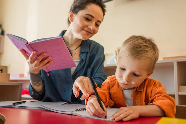 Profesor sonriente señalando con el dedo cerca de niño escribiendo dictado en la escuela montessori - foto de stock
