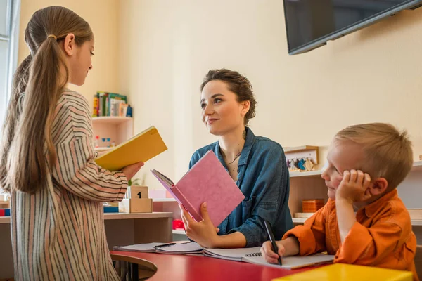 Девочка с книгой, стоящей рядом с учителем и мальчиком, сидящих за столом в классе — стоковое фото