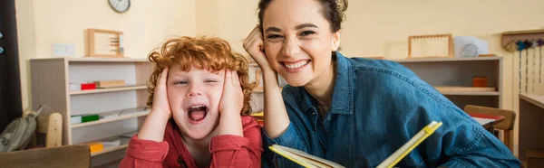 Cheerful teacher holding book near redhead astonished boy, banner — Stock Photo