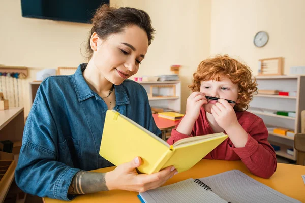 Garçon tenant stylo près du visage tandis que montessori professeur d'école livre de lecture — Photo de stock