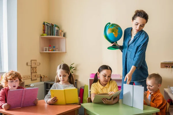 Teacher with globe pointing at book near interracial kids reading in classroom — Stock Photo